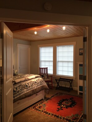 Une chambre à coucher avec un lit, un tapis rouge, un fauteuil à bascule et deux fenêtres équipées de stores.