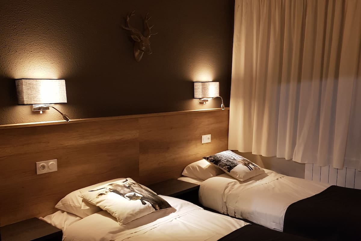 A hotel room with two beds, a wooden headboard, wall-mounted lamps, a mounted deer head, and a window with curtains.