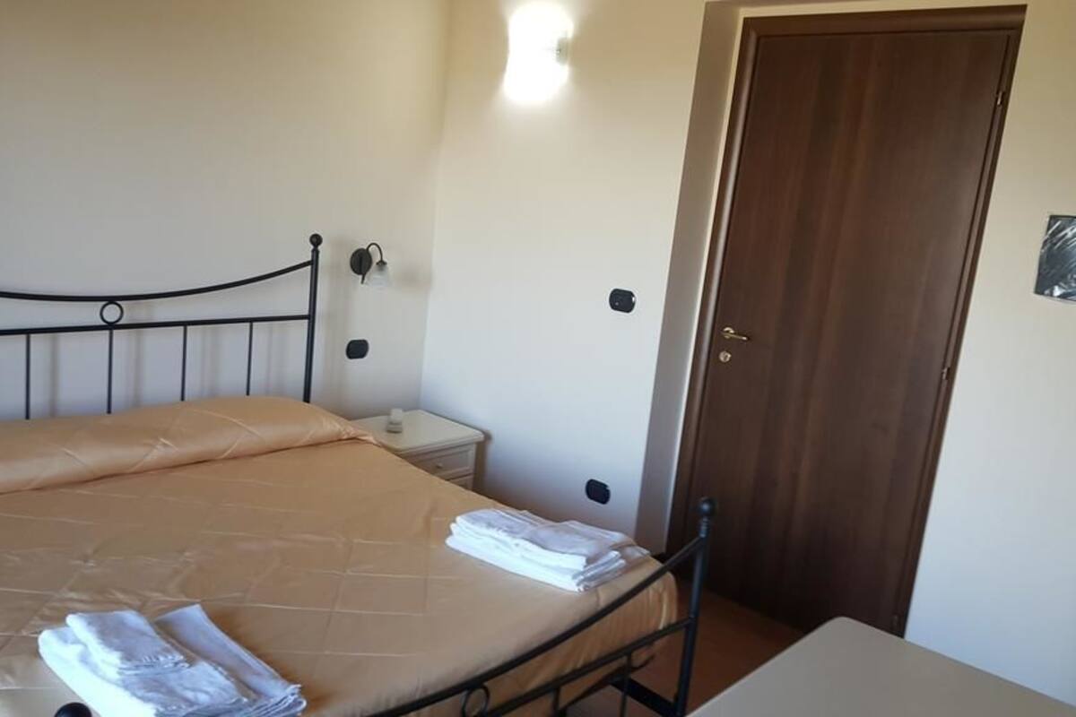Ein Einzelbett mit Kopfteil, ein Nachttisch und eine Nachttischlampe. Eine Holztür und eine Wandleuchte sind ebenfalls vorhanden.