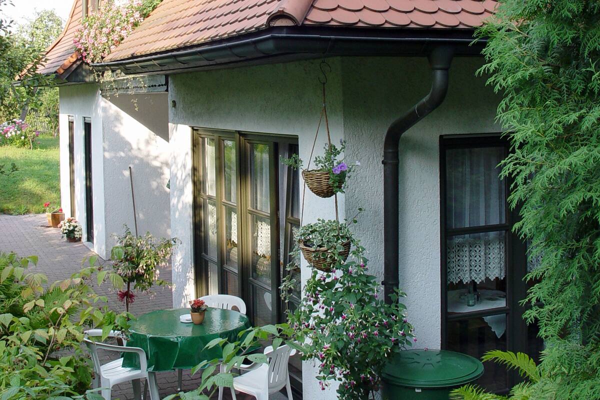 Une maison avec un toit en tuiles, une véranda à l’avant avec une table et des chaises, et des jardinières suspendues remplies de fleurs.
