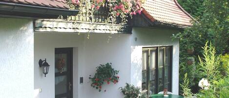 Una casa de dos pisos con un balcón decorado con flores, una banca de madera y una mesa con sillas afuera.