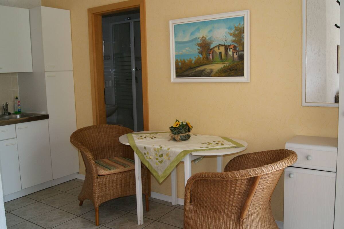 Un petit coin repas avec des chaises en osier, une table agrémentée d’un arrangement floral et un tableau accroché au mur.