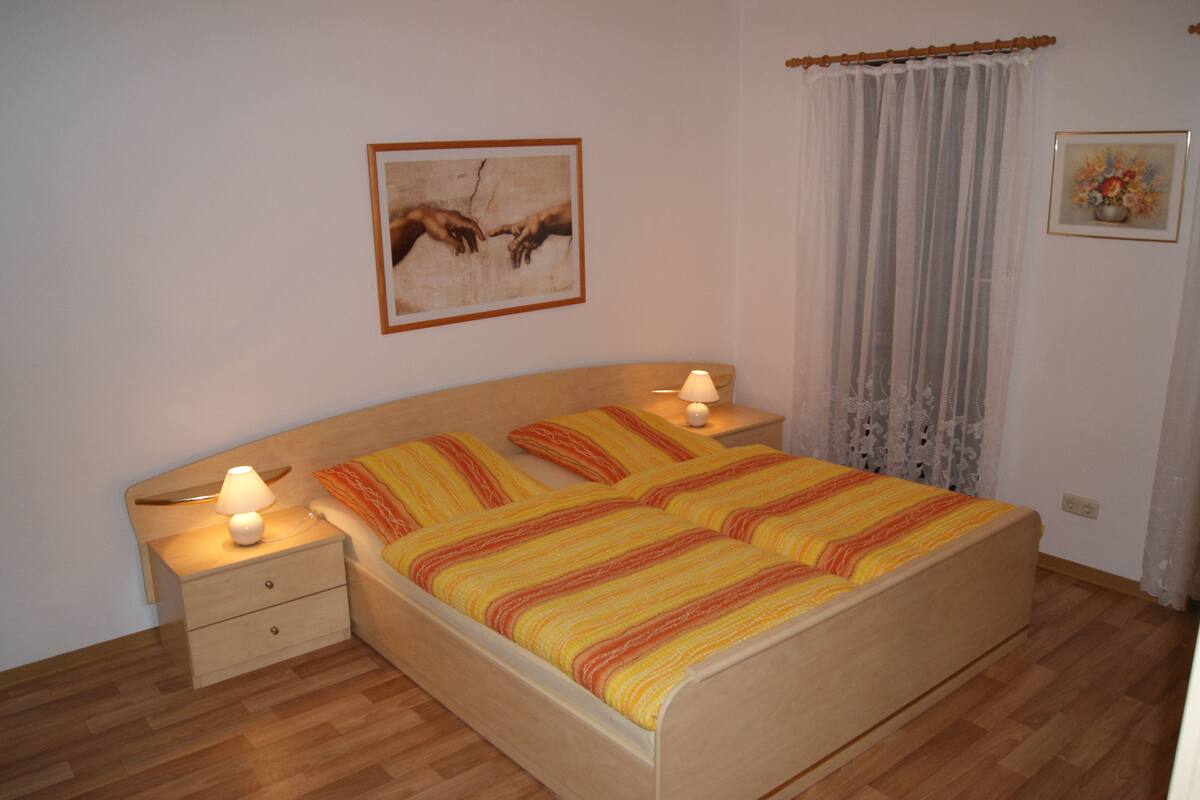 Une chambre à coucher avec un lit, deux tables de chevet, une commode, une lampe, un tableau encadré au mur et une fenêtre avec des rideaux.