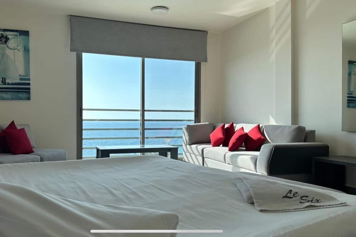 A hotel room with a large bed, two sofas, a small table, and a view of the sea through large windows.