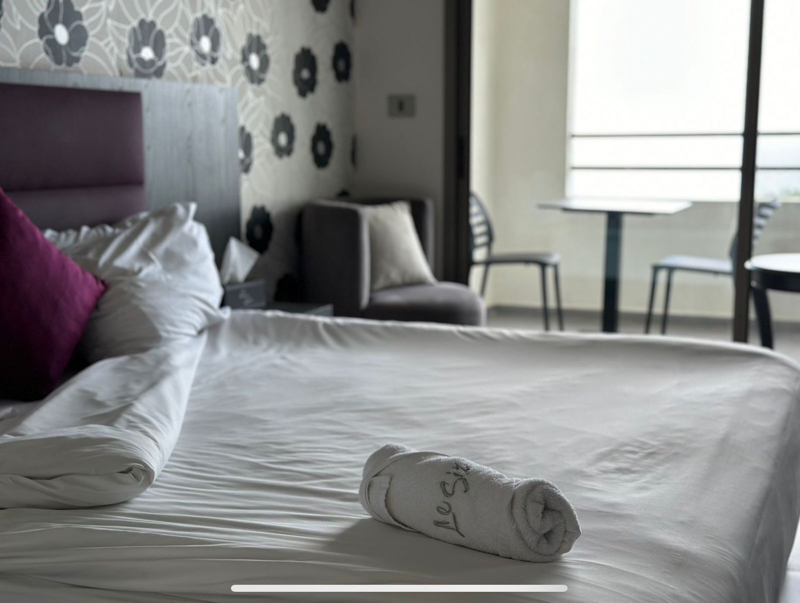 Cama bem arrumada com uma toalha enrolada e um travesseiro bordado com 'Le Site'.