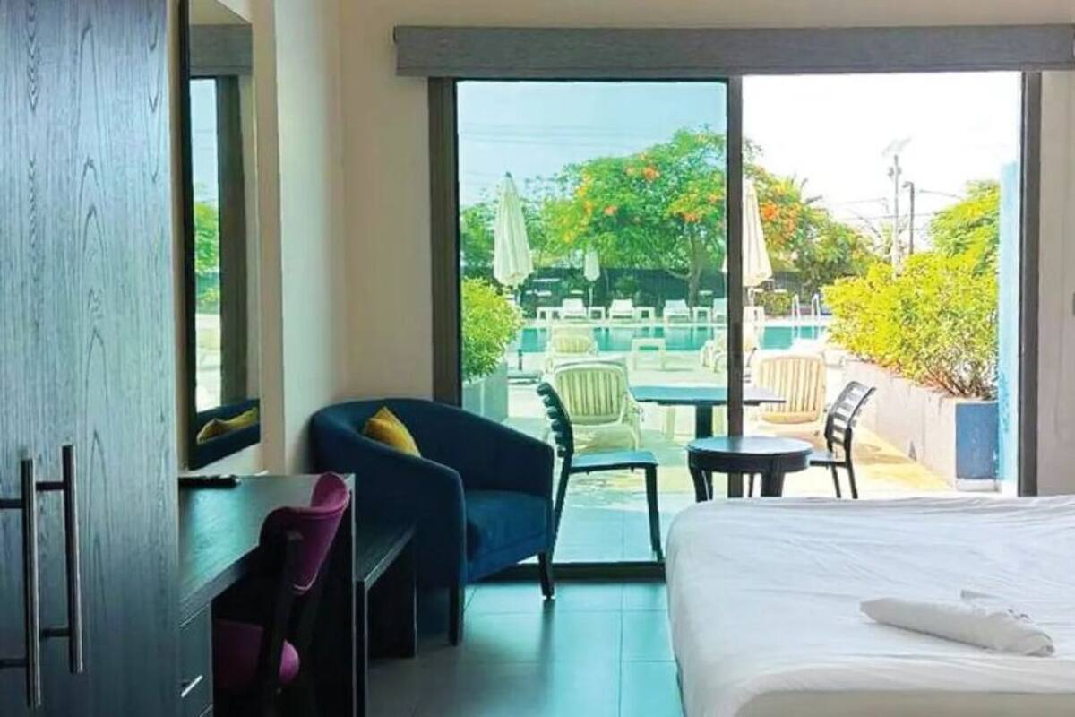 A hotel room with a bed, a desk, a chair, a small table, and a view of an outdoor pool area.