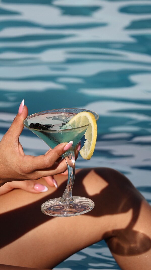 A person holding a cocktail glass with a lemon slice, near a body of water. - Le Six Resort Hotel (Batroun)