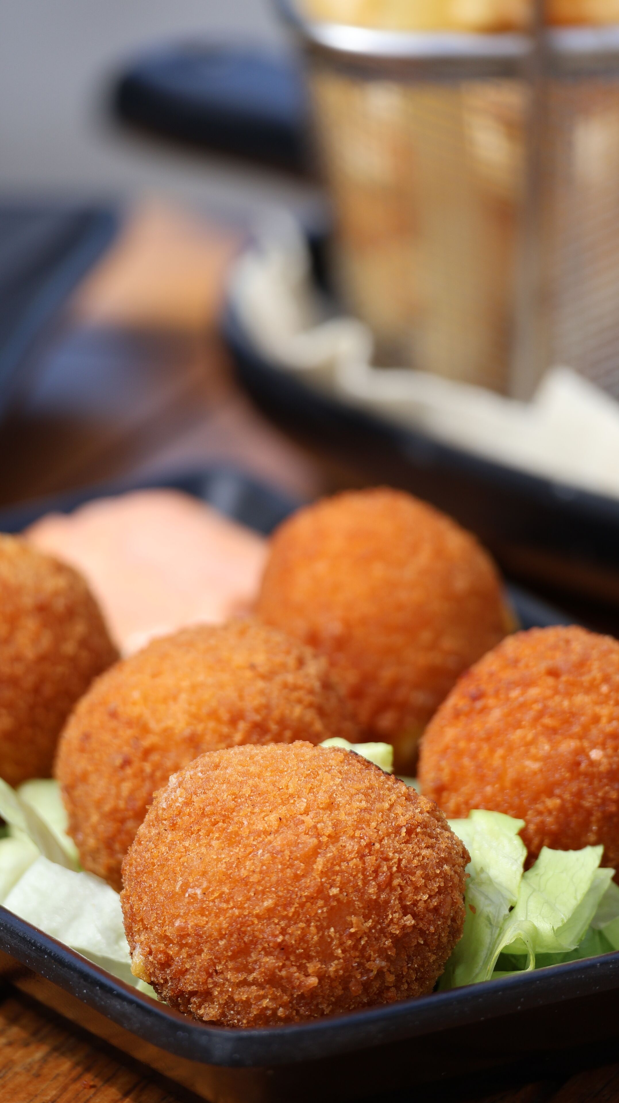 A tray of breaded and fried balls, possibly croquettes, served on a bed of lettuce with a side of sauce.