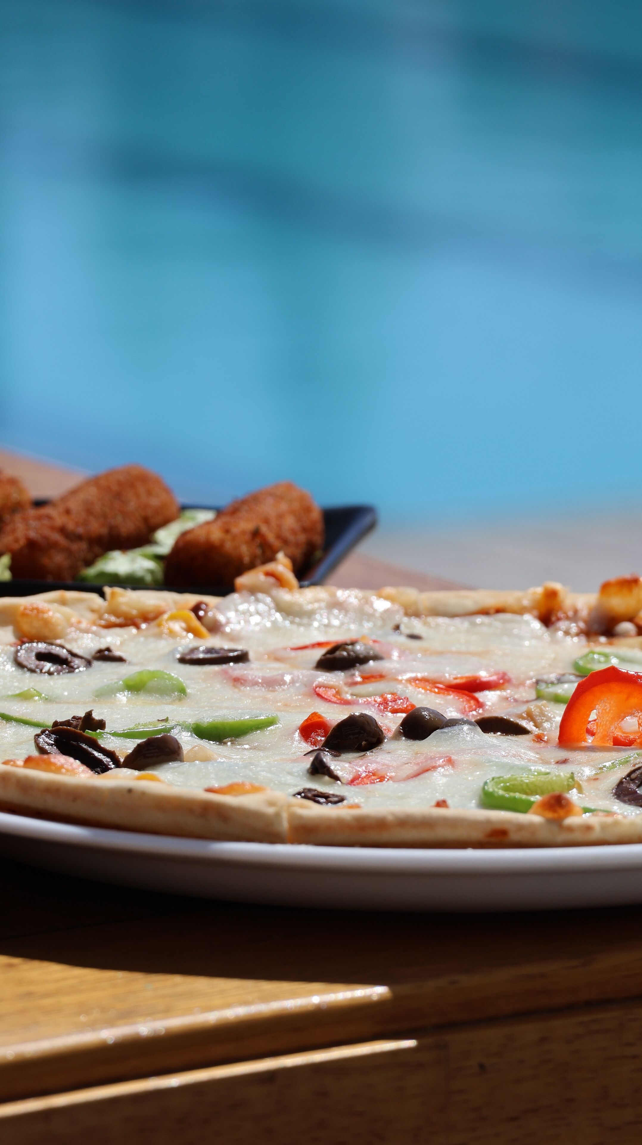A pizza with various toppings on a plate, with a pool in the background.
