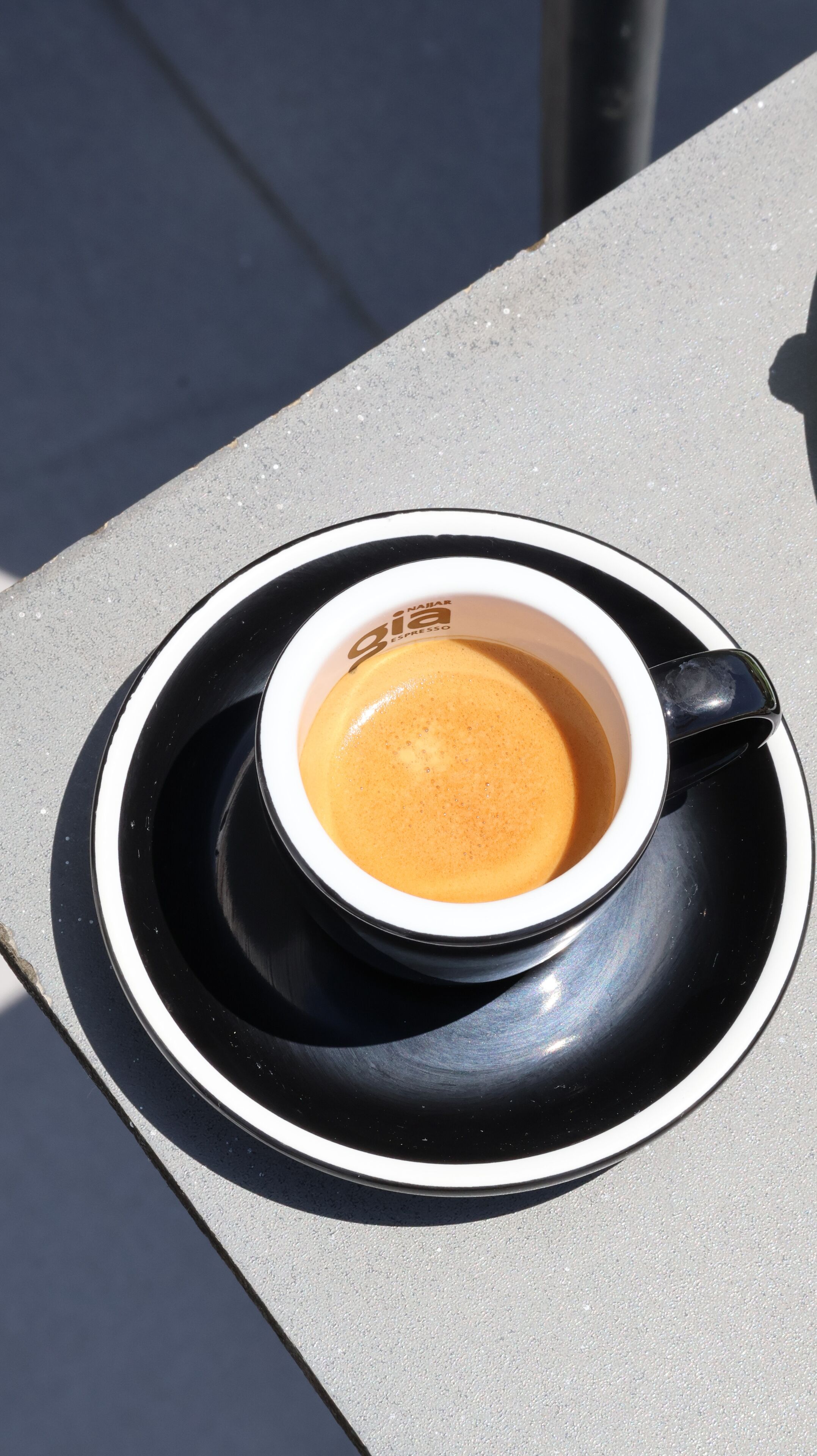 A cup of coffee on a black and white saucer with a logo, placed on a gray surface.