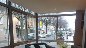 A modern interior with a sofa, chairs, and a small table. Large windows offer a view of the street and buildings outside. - Hotel Nestor (Bourg-les-Valence)