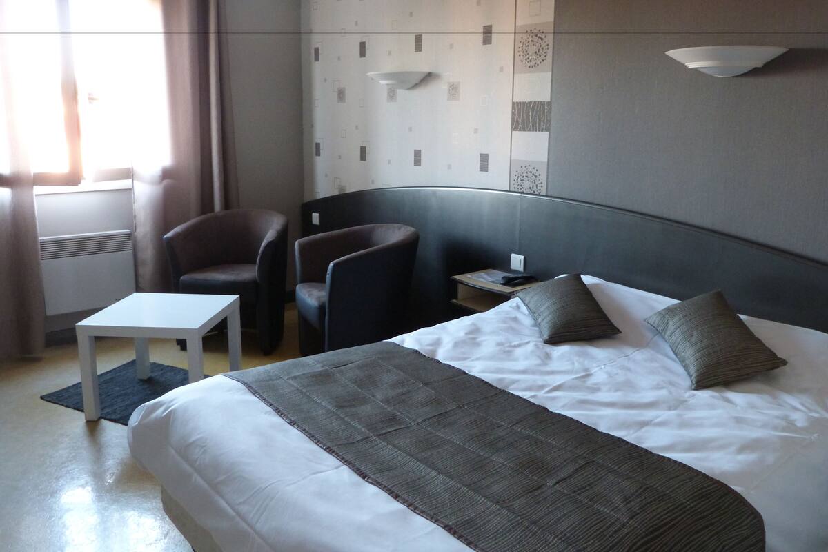 Een hotelkamer met een groot bed, twee fauteuils, een klein tafeltje, een nachtkastje en een wandlamp.