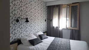 A modern hotel room with a geometric patterned wall, a large bed with white and gray bedding, two black wall sconces, and a window with blinds. - Hotel Nestor (Bourg-les-Valence)