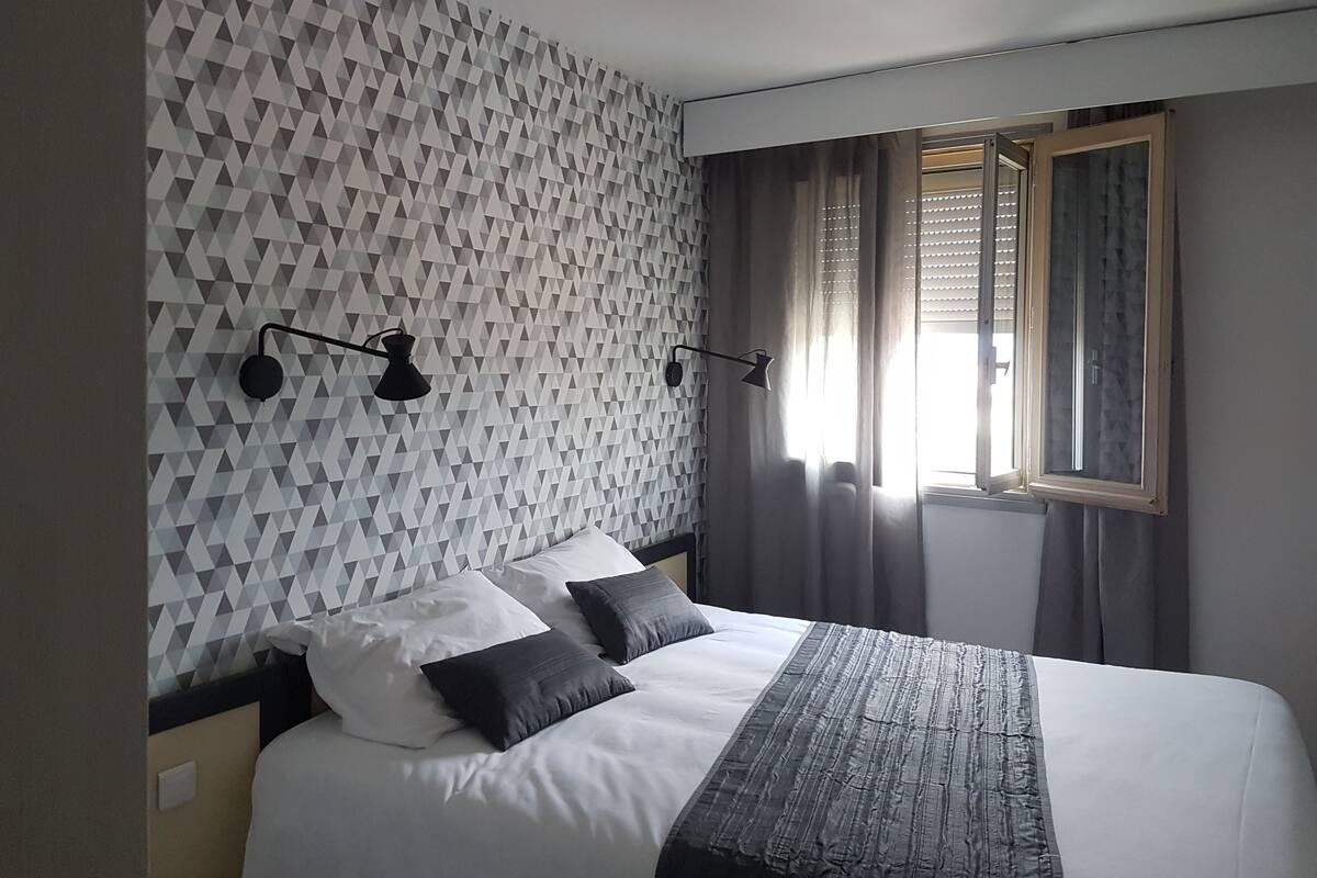 Een moderne hotelkamer met een wand met geometrisch patroon, een groot bed met wit en grijs beddengoed, twee zwarte wandlampen en een raam met jaloezieën.