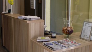 A reception desk with a wooden finish, a vase with yellow flowers, a clock, and a glass bowl with candies. - Hotel Nestor (Bourg-les-Valence)