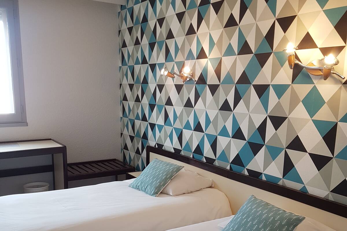 Een hotelkamer met twee bedden, een wand met een geometrisch patroon en een nachtkastje met een lamp.