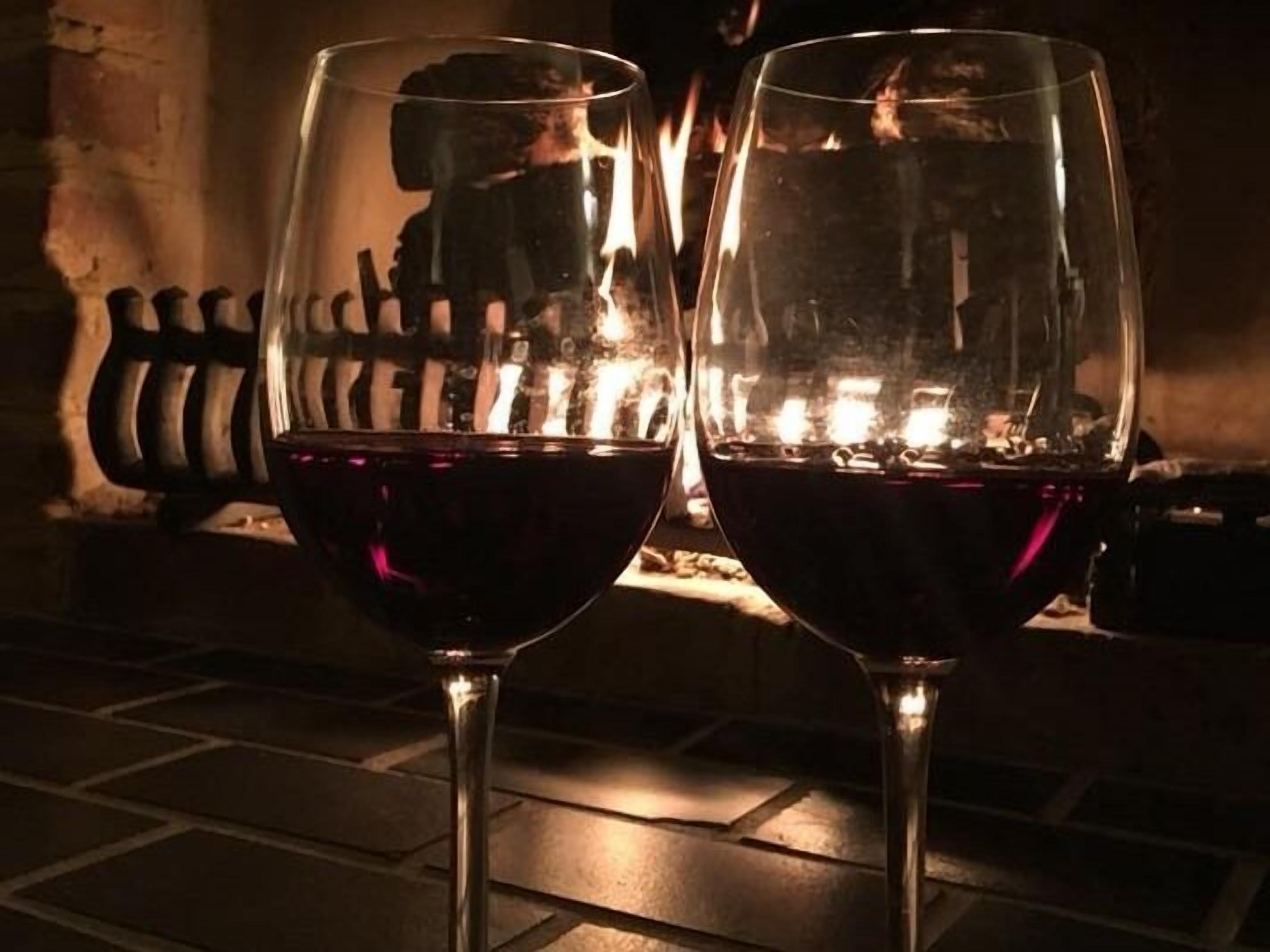 Two wine glasses filled with red wine, placed on a stone floor with a fire in the background.