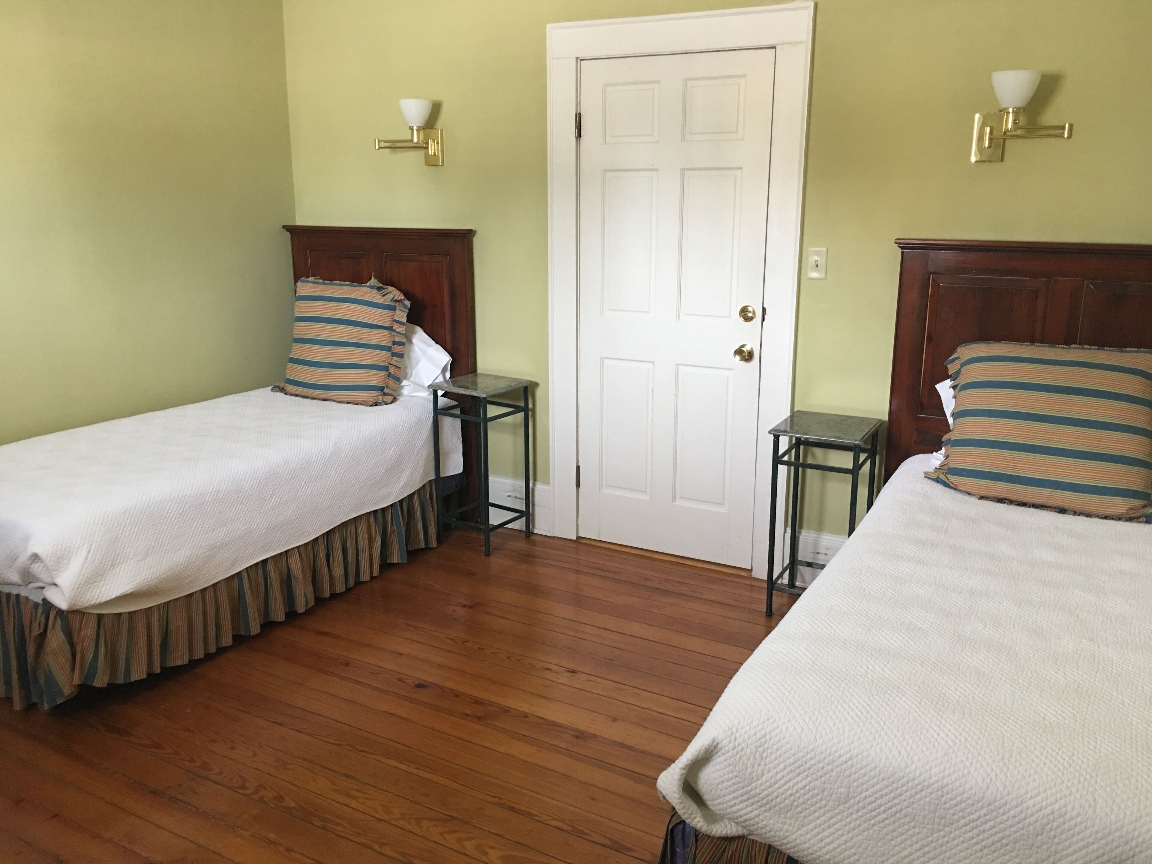 Quarto com duas camas de solteiro, piso de madeira, porta branca e luminárias fixadas na parede.