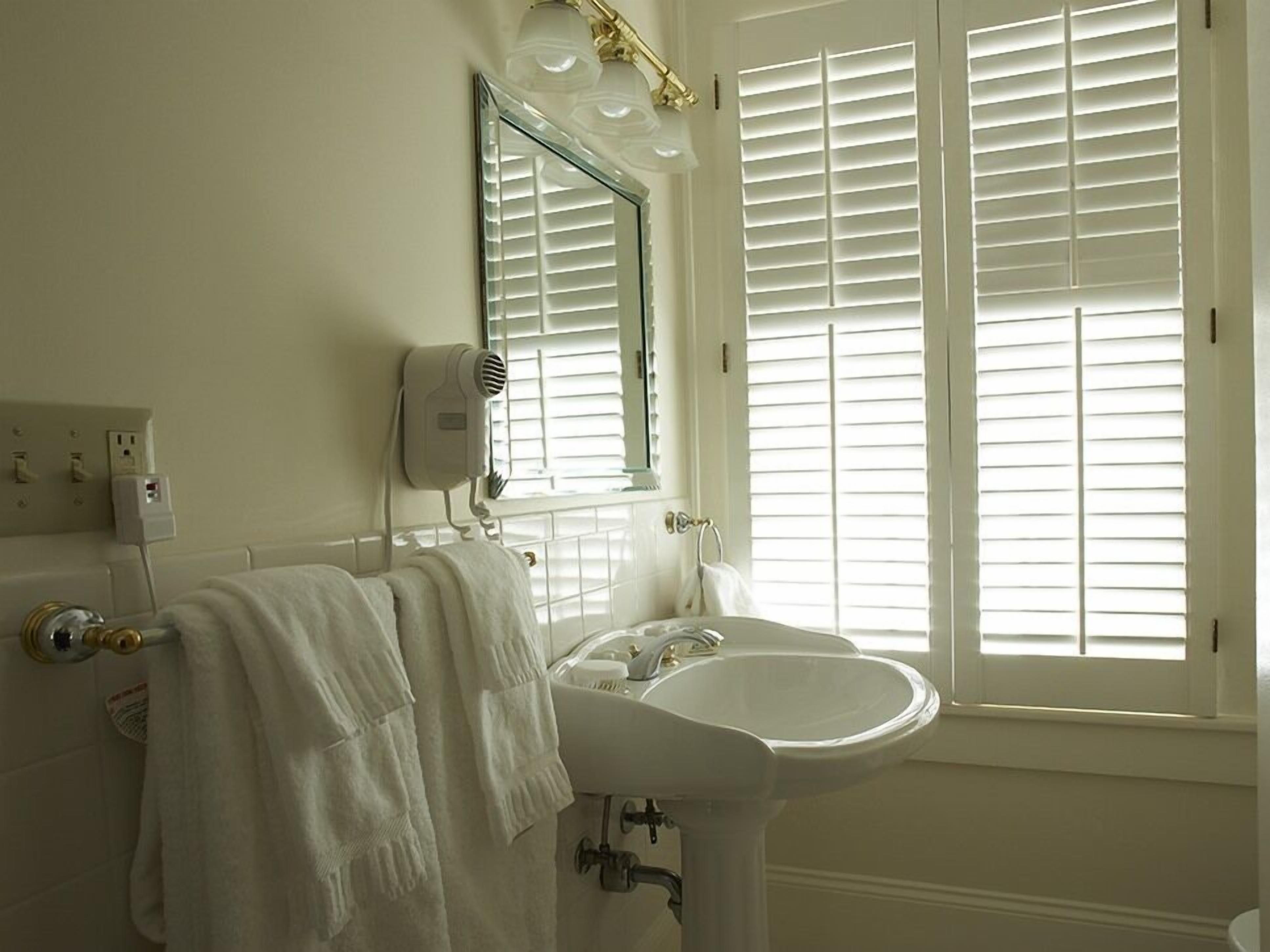 Une salle de bain avec un lavabo blanc, un miroir avec éclairage intégré, un sèche-cheveux et une fenêtre avec stores.
