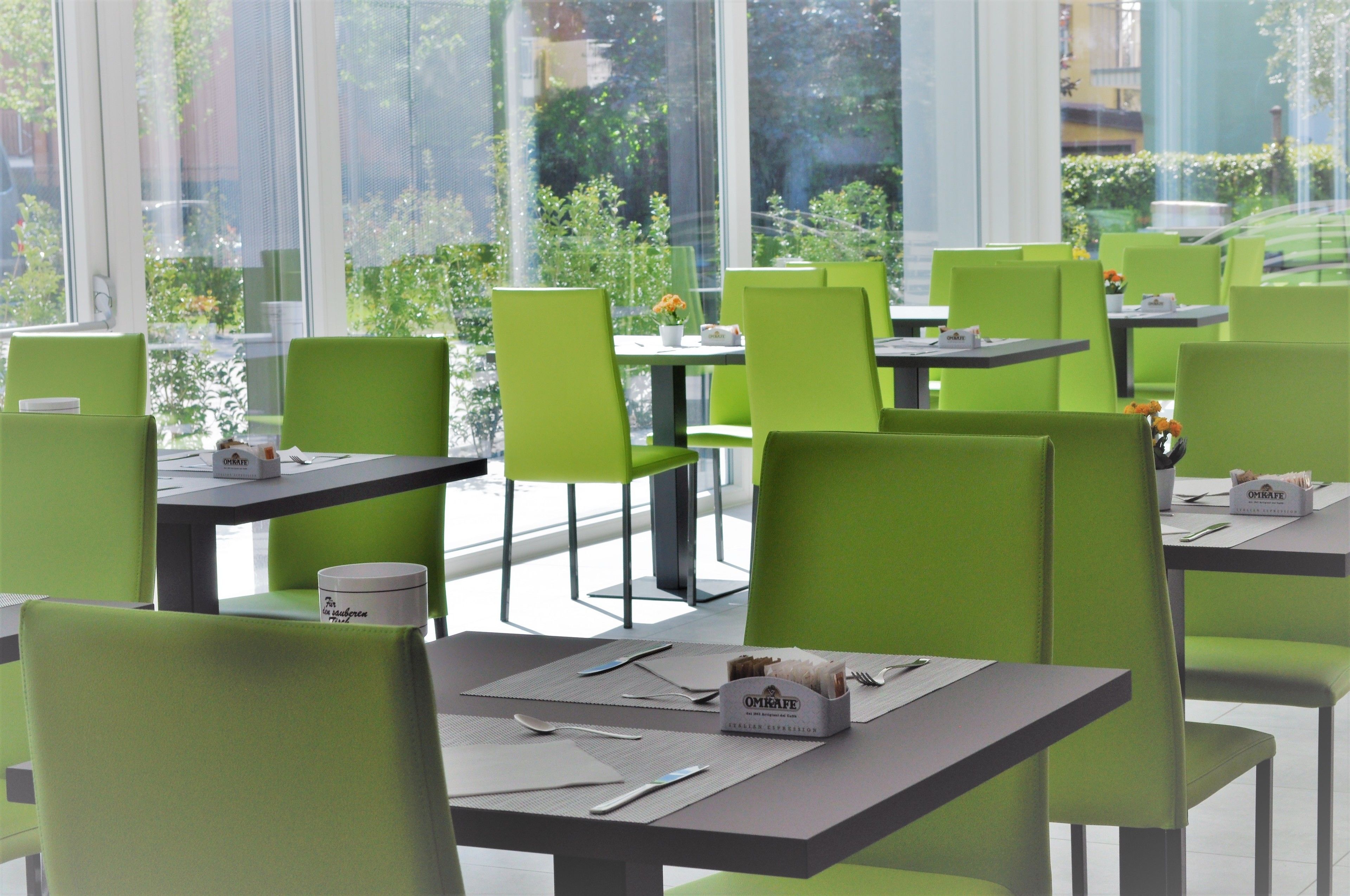 A modern cafe with green chairs and tables, outdoor seating, and large windows offering views of greenery and buildings.