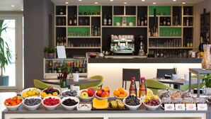 A breakfast buffet with various fruits and beverages, a bar area with shelves of bottles, and a green plant in the background.