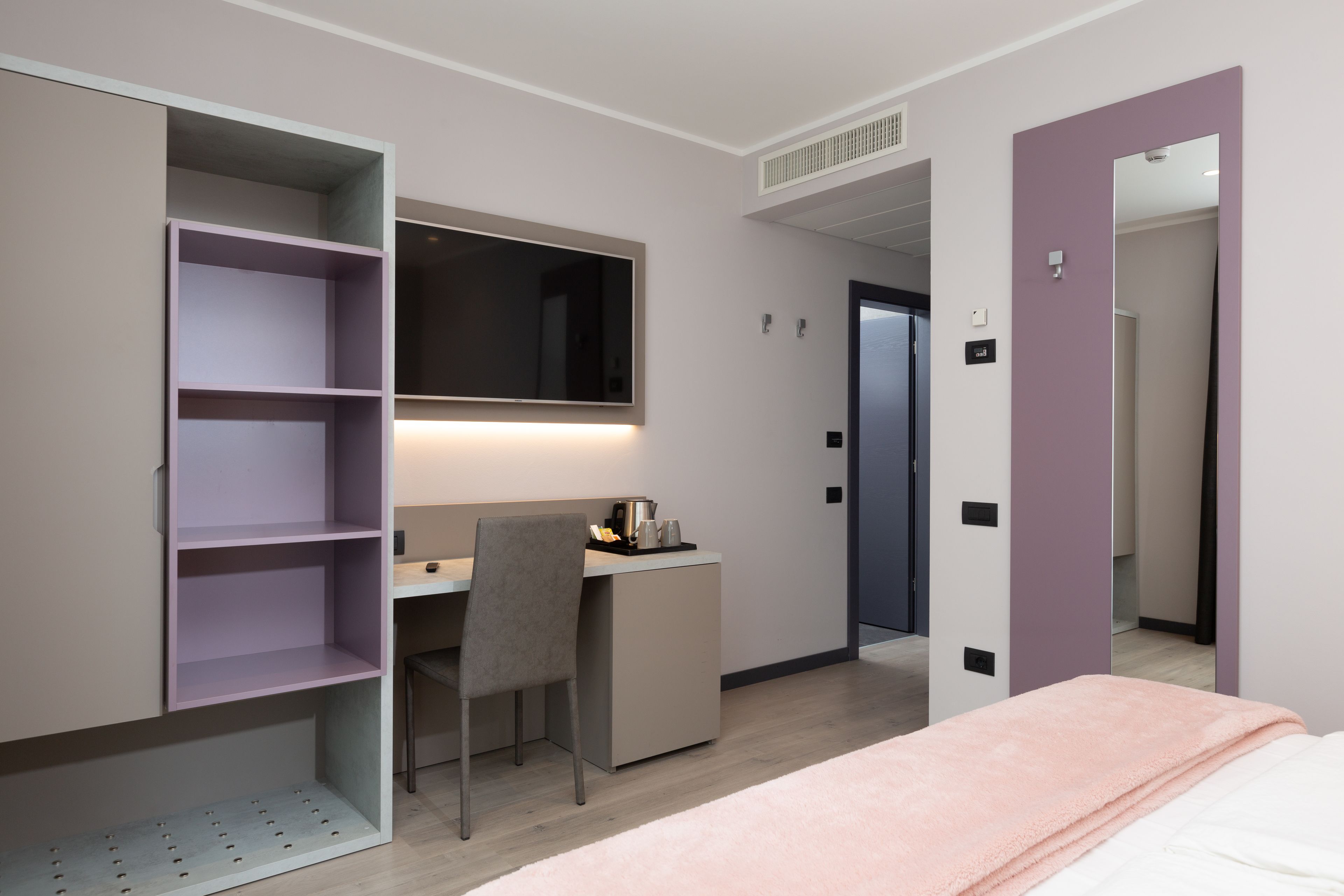 Una habitación de hotel moderna con cama, escritorio con silla, armario con estantes y espejo.