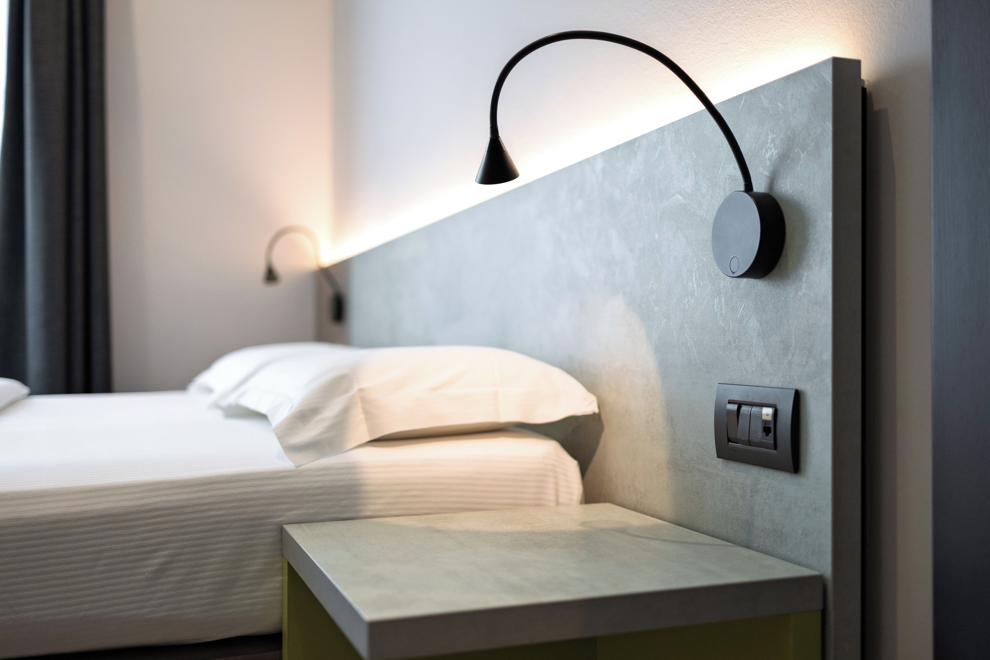 A modern hotel room with a neatly made bed, a bedside table, and a wall-mounted light fixture.