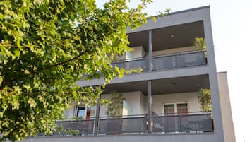 A multi-story building with balconies, large windows, and a tree in the foreground.