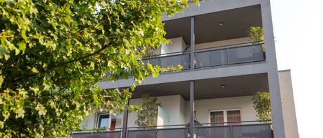 A multi-story building with balconies, large windows, and a tree in the foreground.