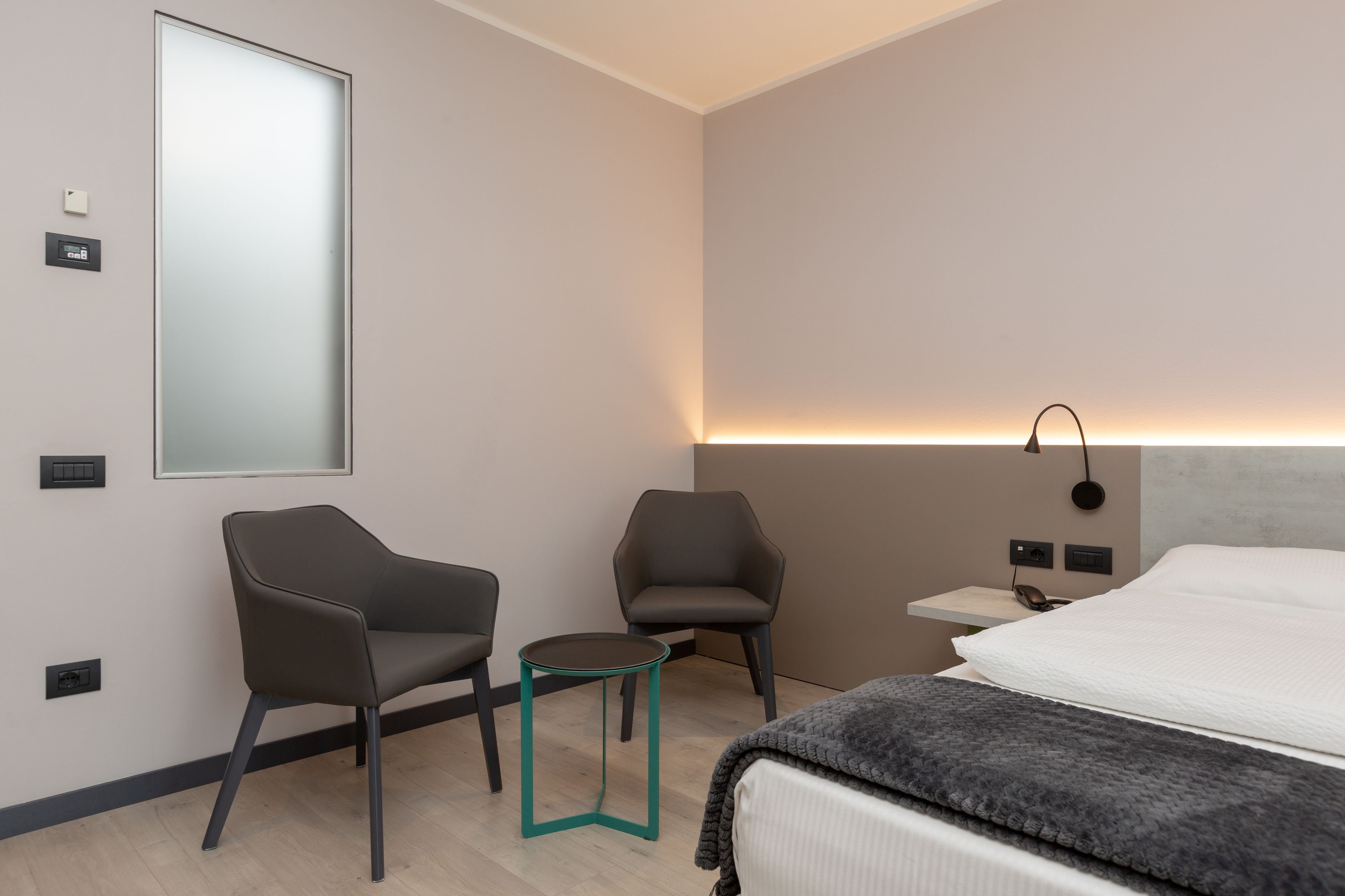 Ein modernes Hotelzimmer mit einem großen Bett, zwei Sesseln, einem kleinen Tisch und einem Nachttisch mit Lampe.