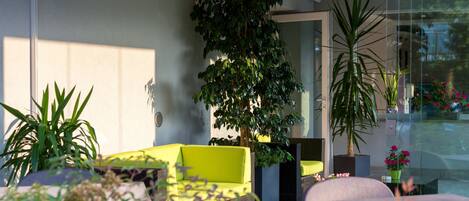 A modern outdoor seating area with a bright yellow sofa, a large potted plant, and a glass door leading to a garden.