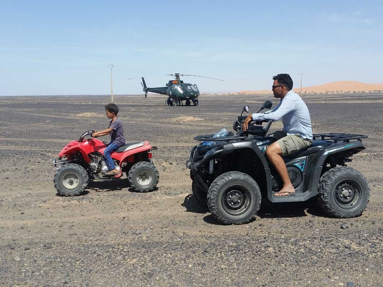 A desert landscape with a helicopter in the background, a red ATV, and a black ATV.