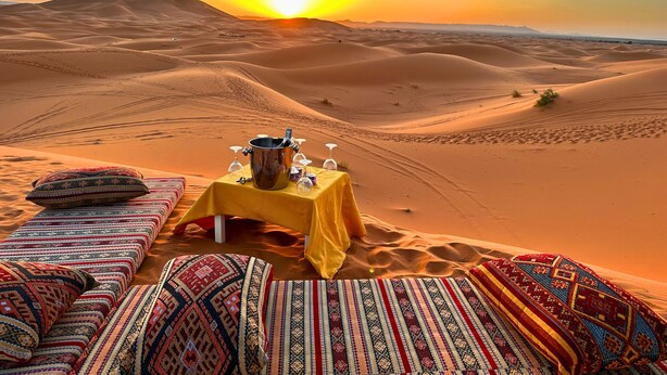 A desert scene with a table set for a meal, surrounded by colorful cushions and a sunset in the background. - DWO Golden Camp Merzouga (Taouz)