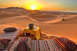 A desert scene with a table set for a meal, surrounded by colorful cushions and a sunset in the background. - DWO Golden Camp Merzouga (Taouz)