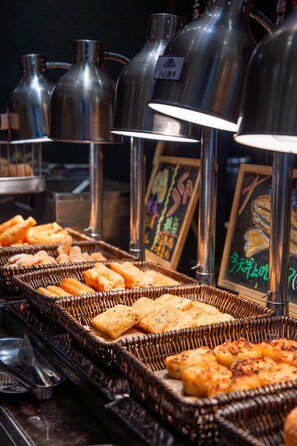 A buffet with various food items, including bread and pastries, under metal serving stations with lights.