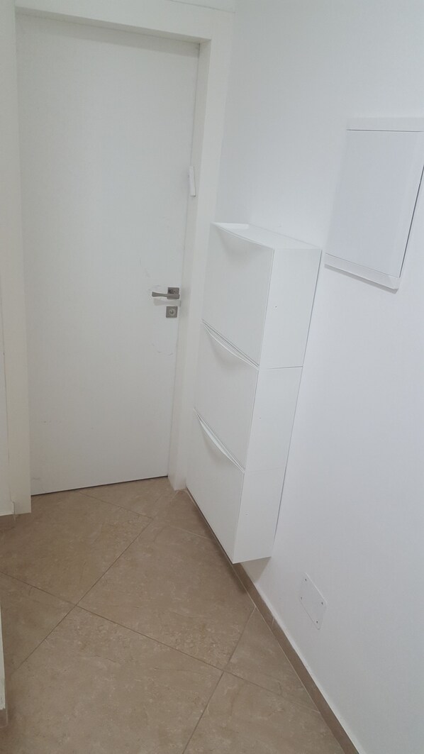 Een kamer met een witte deur, een betegelde vloer en een witte muur met een klein plankje.