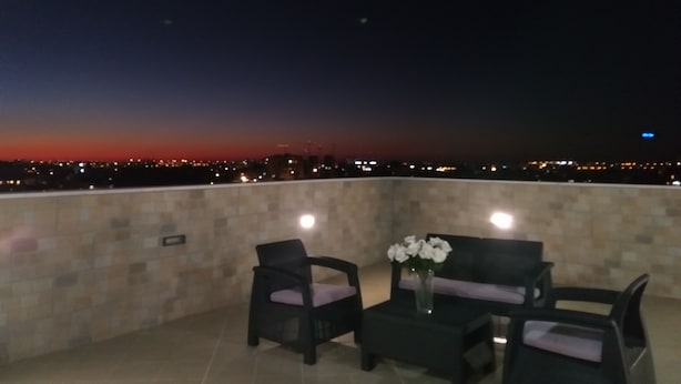 Un balcon donnant sur une vue panoramique de la ville au crépuscule. On y trouve deux chaises et une petite table sur laquelle est posé un vase de fleurs.