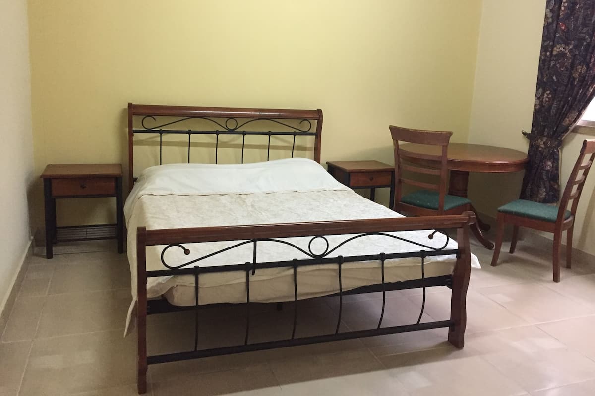 A single bed with a wooden frame and metal headboard, flanked by two nightstands and a wooden chair with a cushion.
