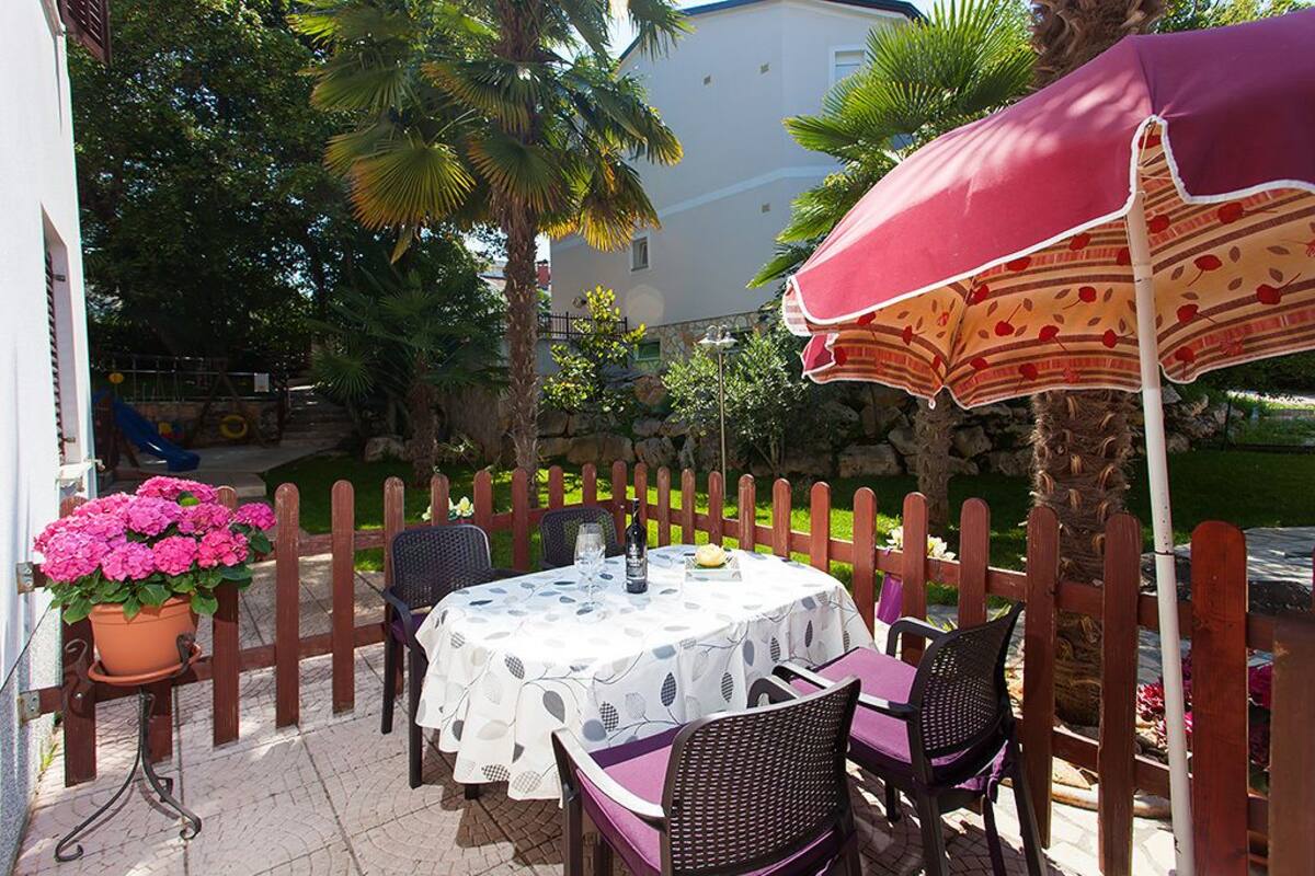 Pátio externo com mesa posta para dois, rodeado por cerca de madeira, e um grande guarda-sol oferecendo sombra.