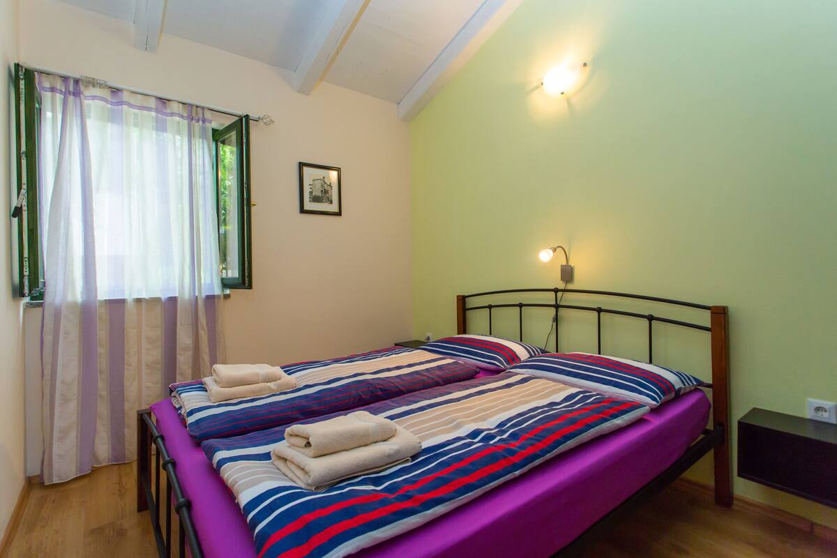 Uma cama de casal com roupa de cama listrada, uma mesinha de cabeceira, uma luminária fixa na parede e uma janela com cortinas.