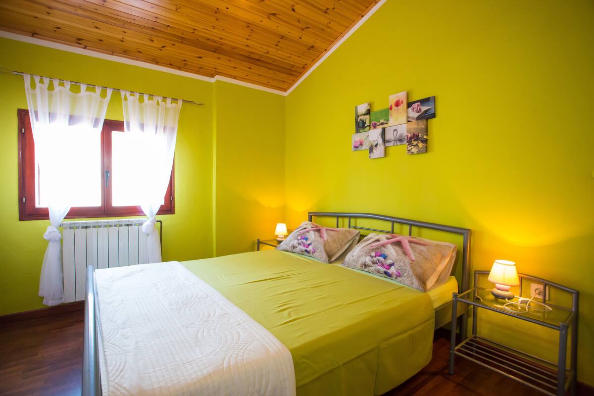 Quarto com cama, mesa de cabeceira com abajur, janela com cortinas e parede com quadros.