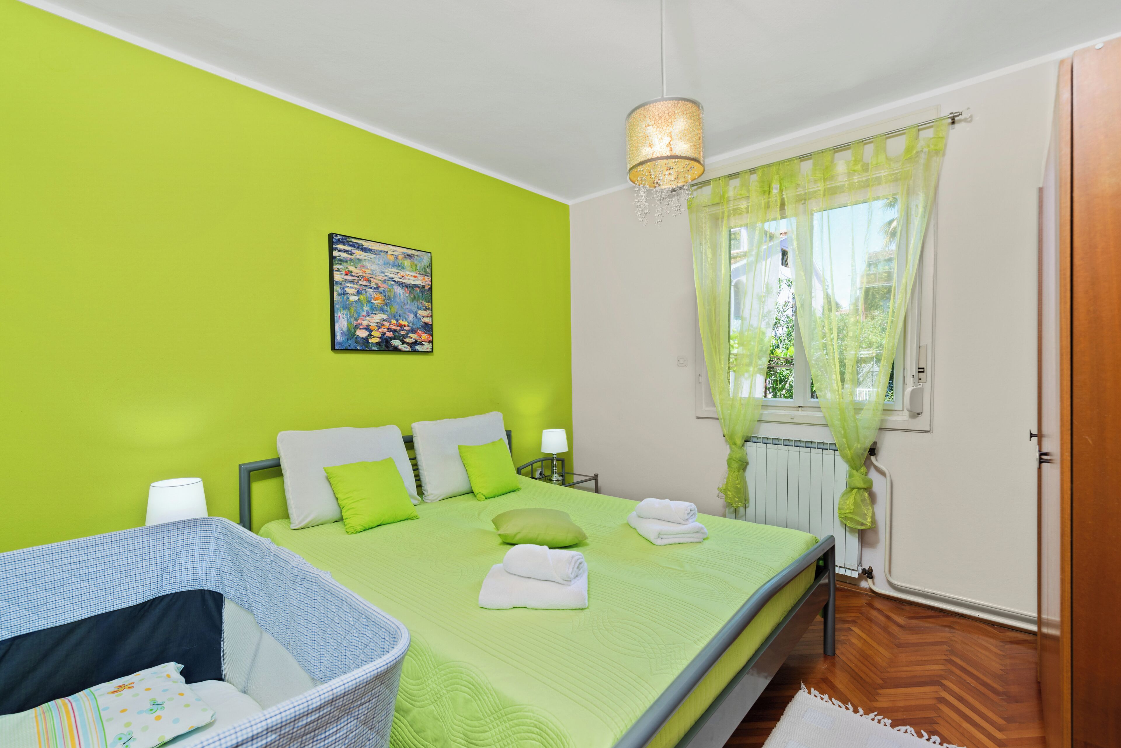 A bedroom with a green wall, a bed with pillows, a bedside table, a lamp, a window with curtains, and a basket.