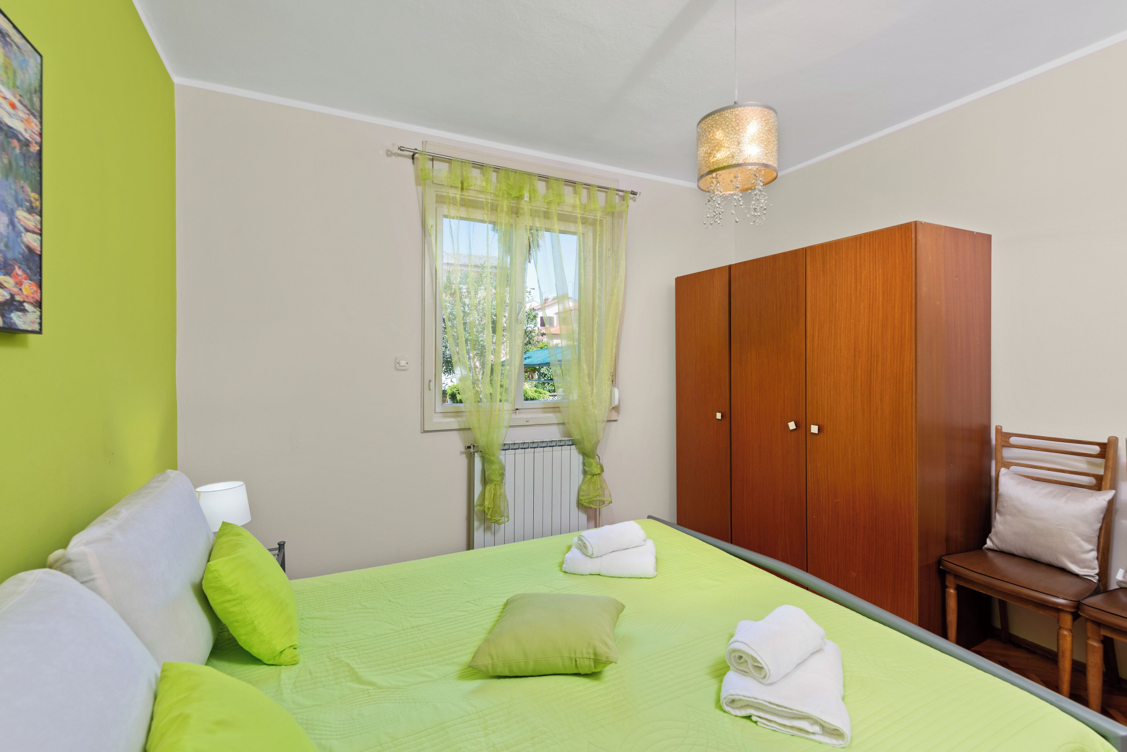 Un dormitorio con cama, ropa de cama verde, un armario de madera, una silla y una ventana con cortinas traslúcidas.