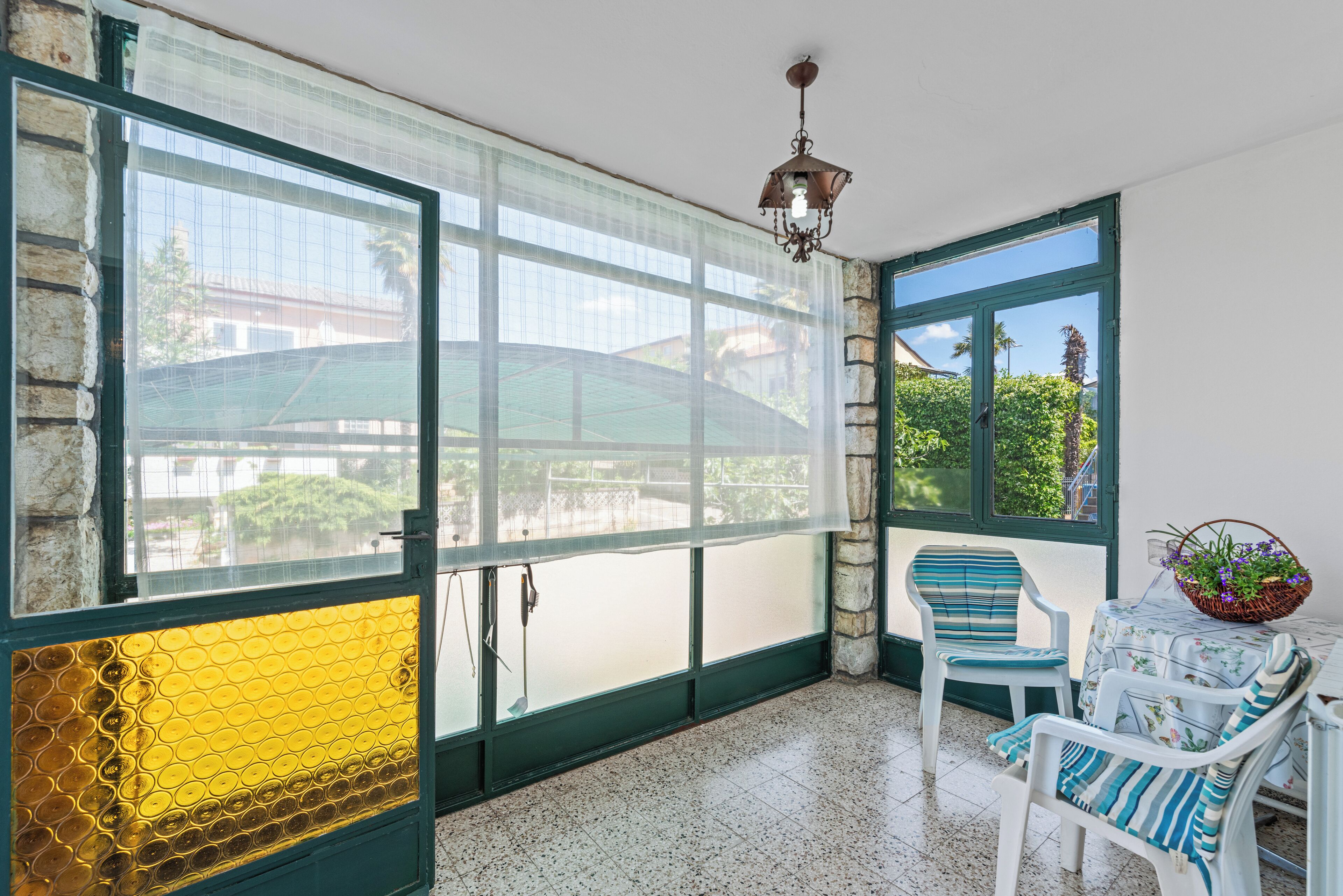 A room with large windows, a chandelier, a table with chairs, and a view of greenery outside.