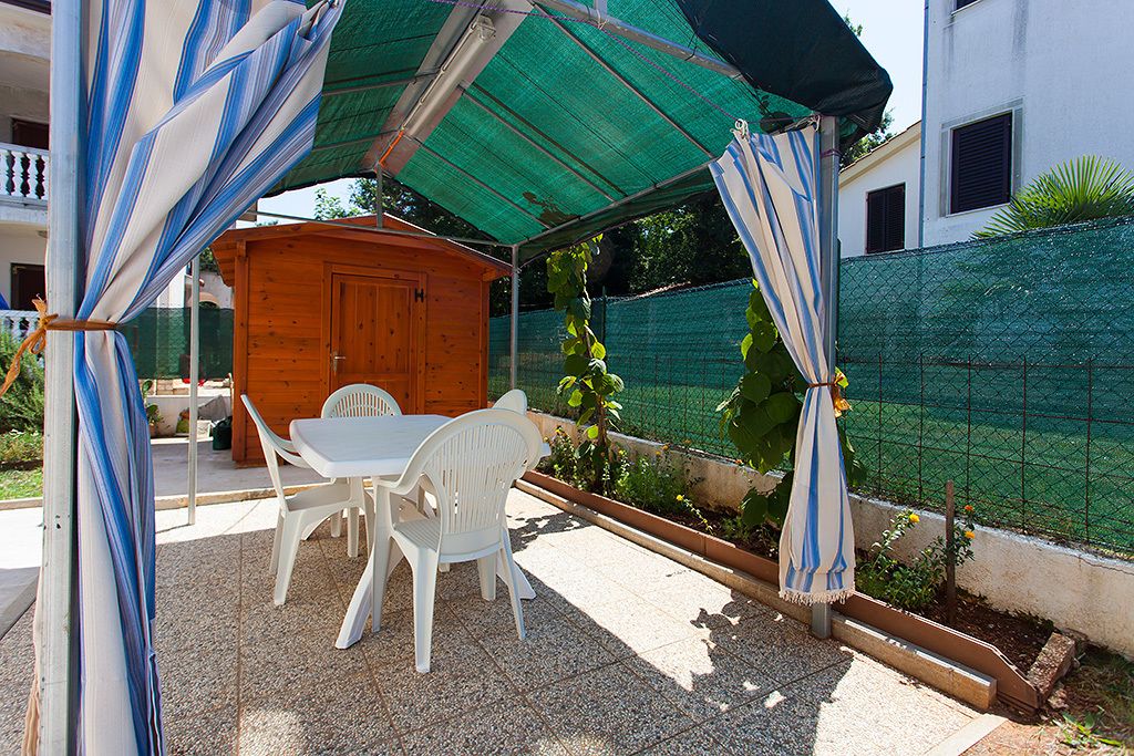 Ein Patio mit einem Holztisch und weißen Plastikstühlen unter einem gestreiften Markisenvorzelt.