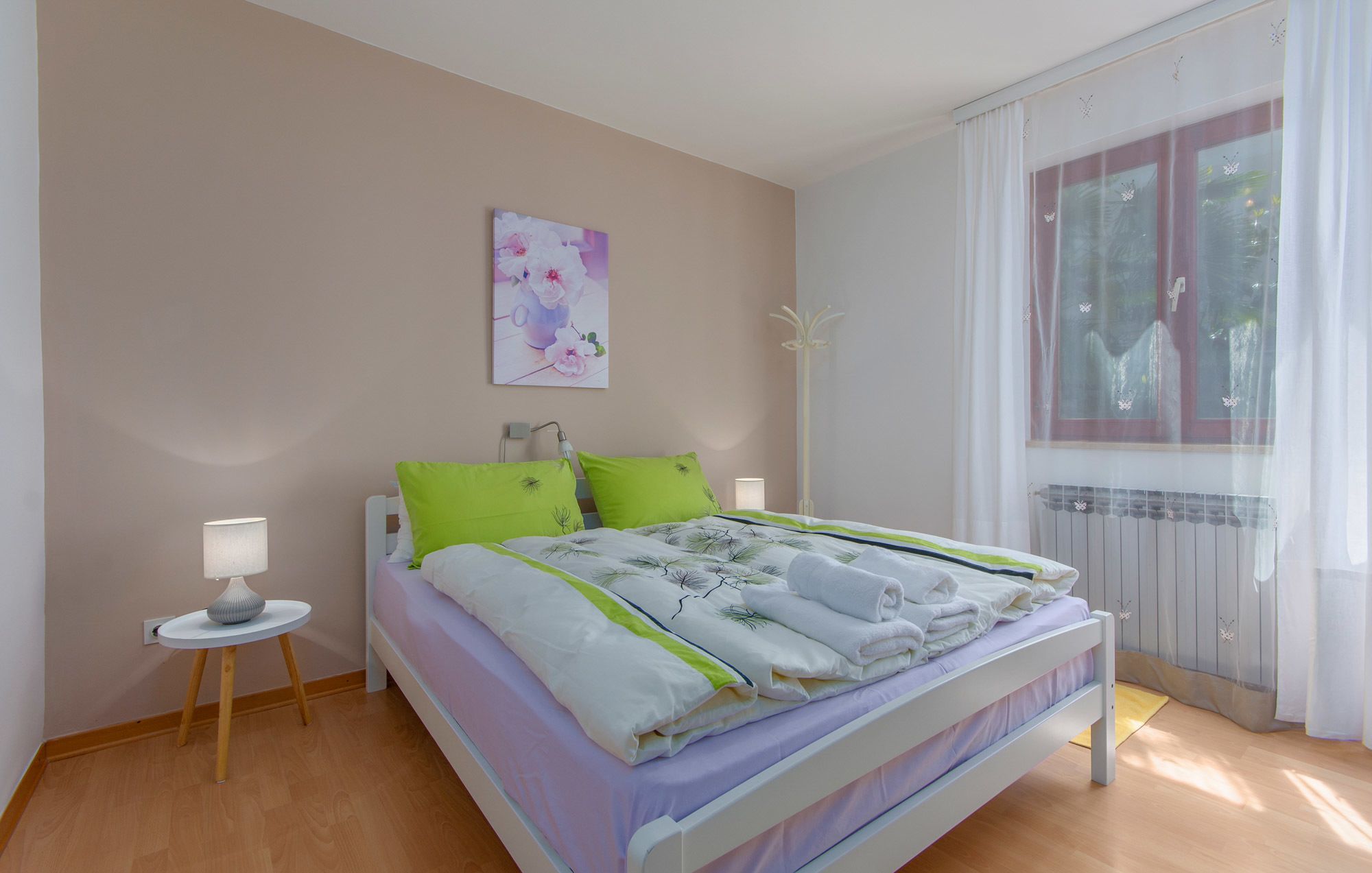 A bedroom with a bed, two bedside tables, a window with curtains, a radiator, and a wall-mounted picture.