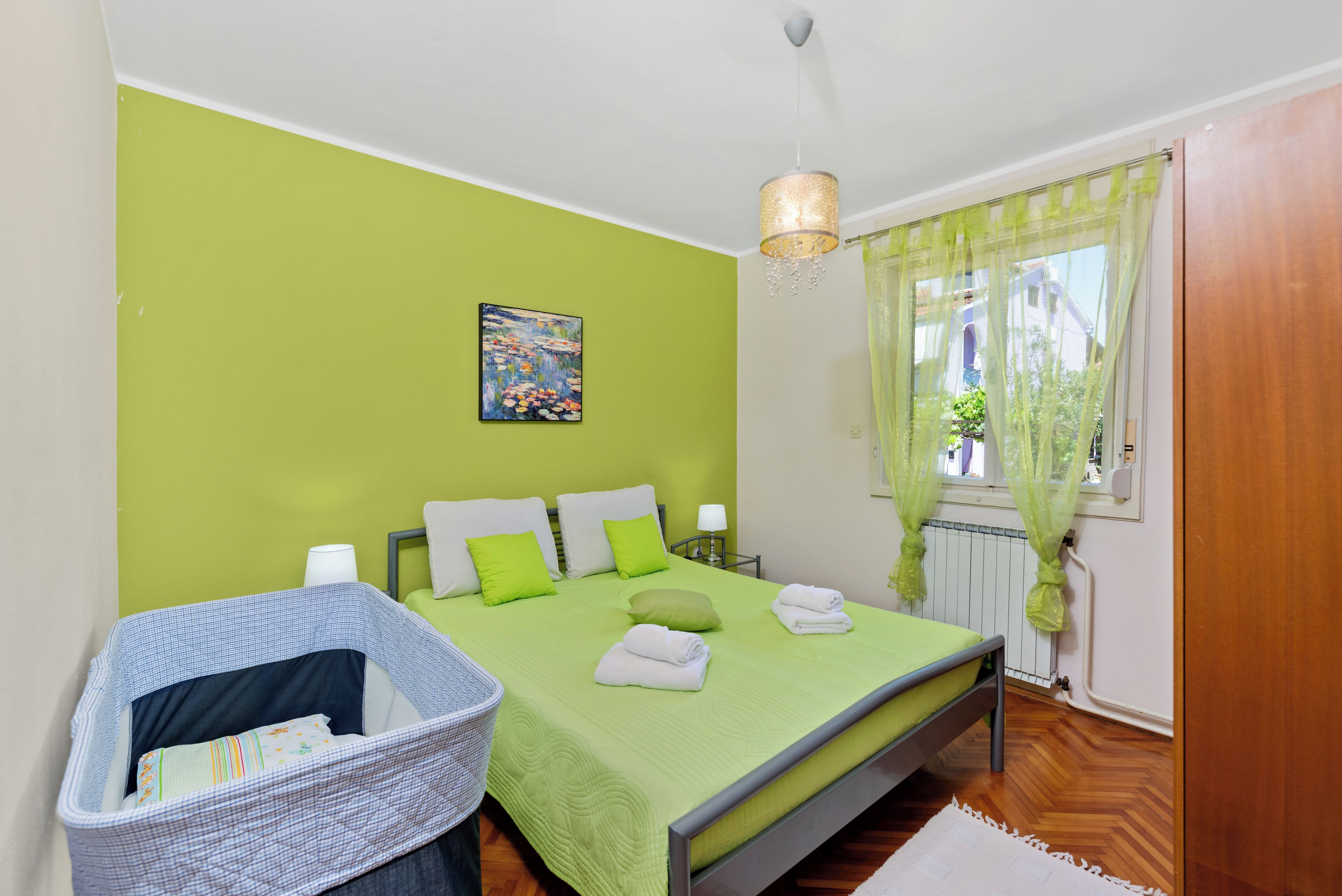 A bedroom with a green wall, a bed with white and green bedding, a bedside table with a lamp, and a window with sheer curtains.