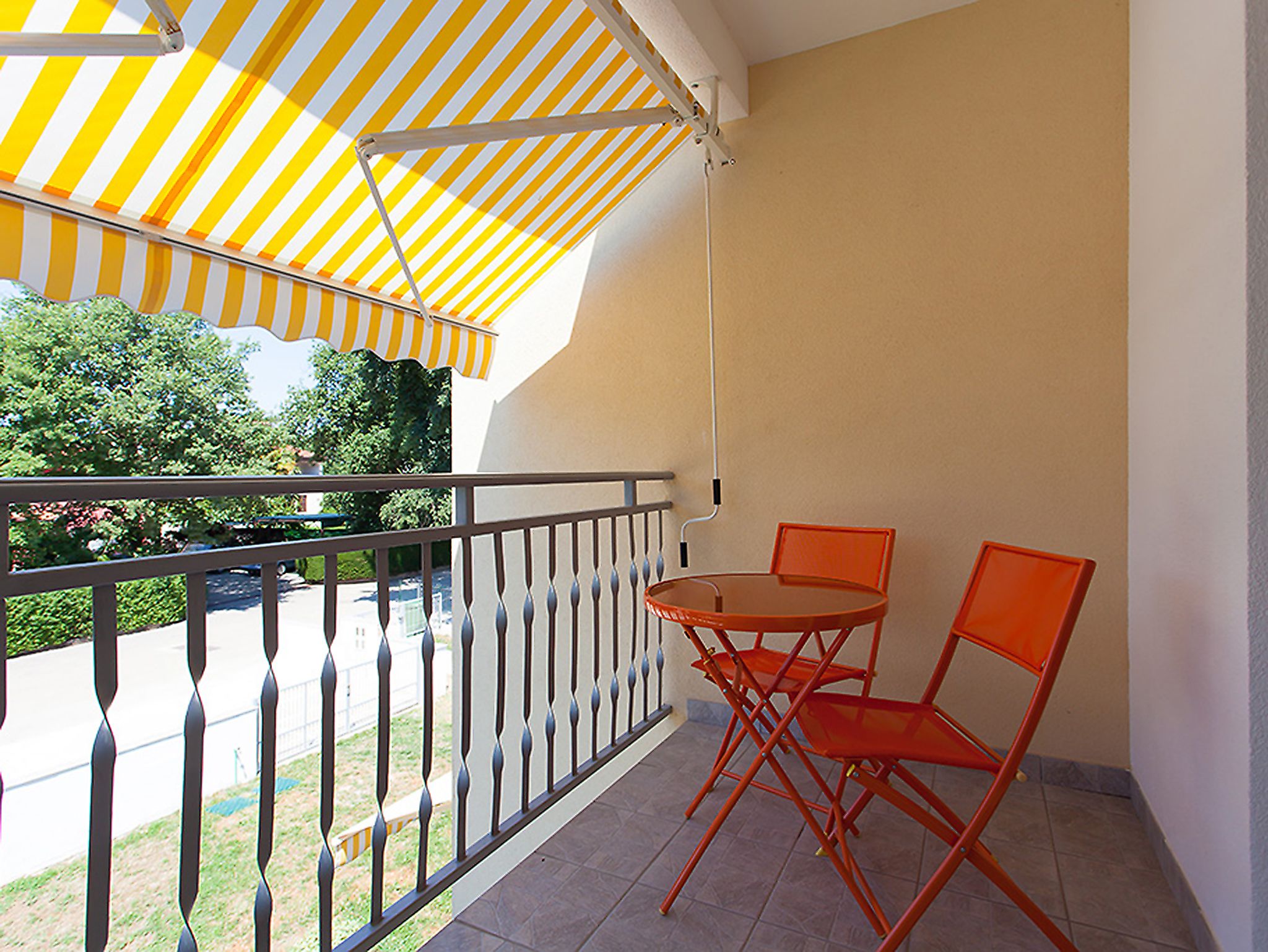 Uma varanda com toldo listrado em amarelo e branco, duas cadeiras laranja e uma pequena mesa redonda.