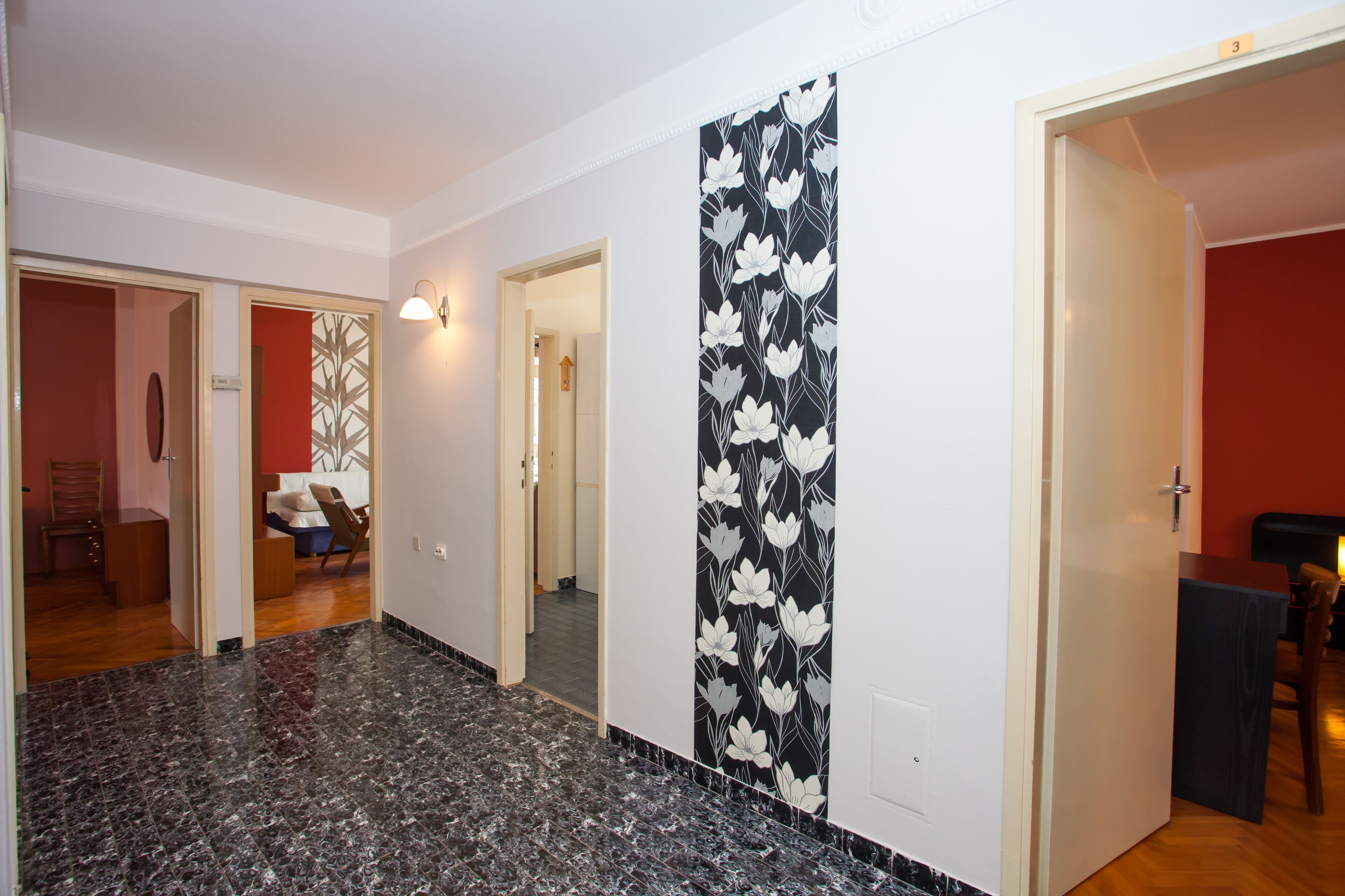 A hallway with a black and white floral wall design, a marble floor, and a door leading to a room with a bed and a chair.