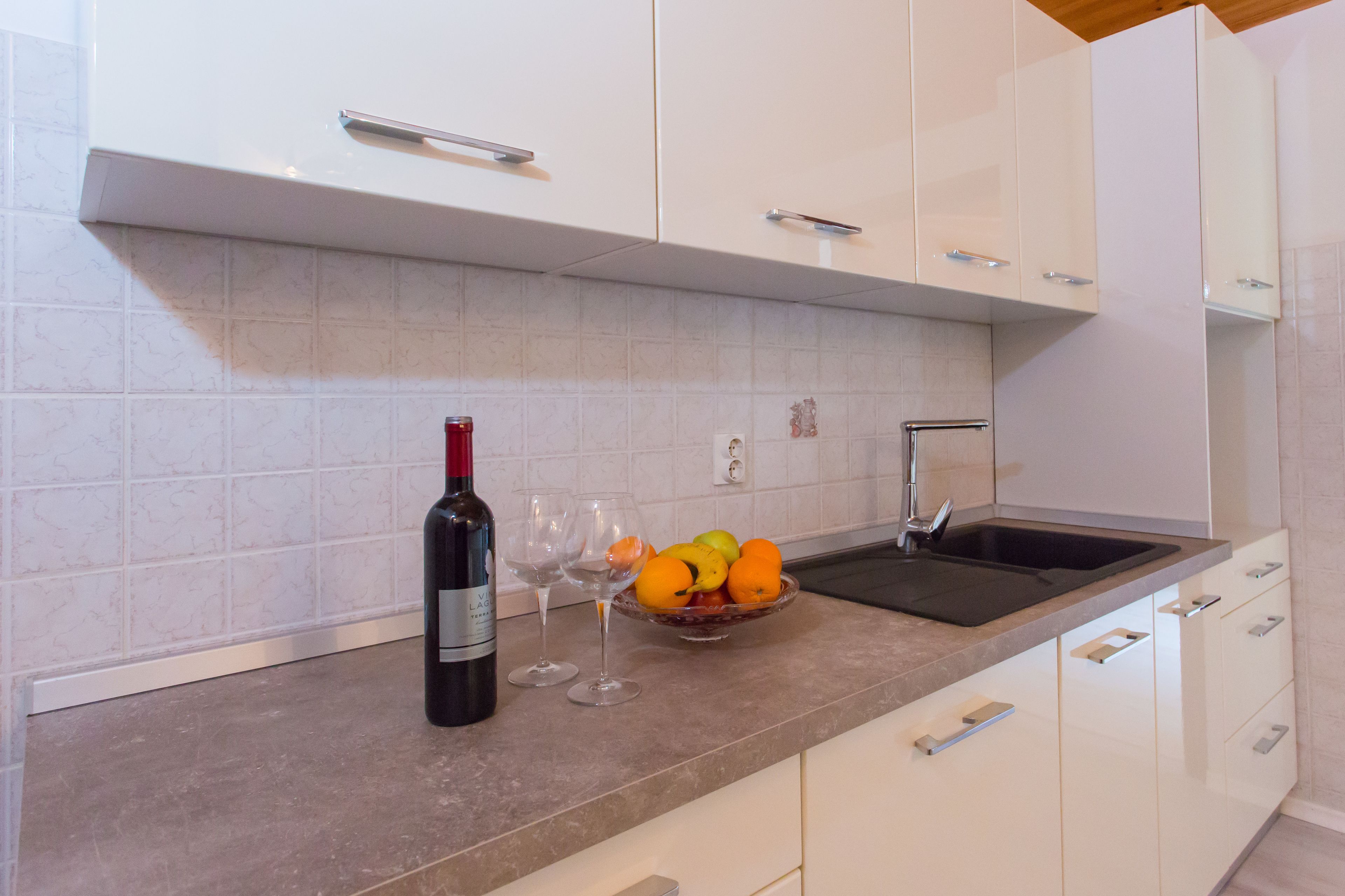 A kitchen with white cabinets, a granite countertop, a wine bottle, two glasses, and a bowl of fruit.